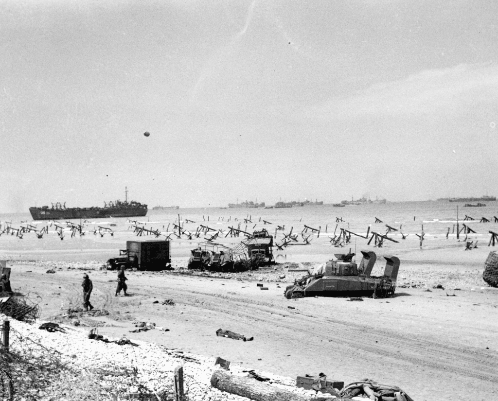Omaha Beach délután a D-napon, elesettekkel, roncs járművekkel, elakadt Sherman harckocsival és parti akadályokkal.